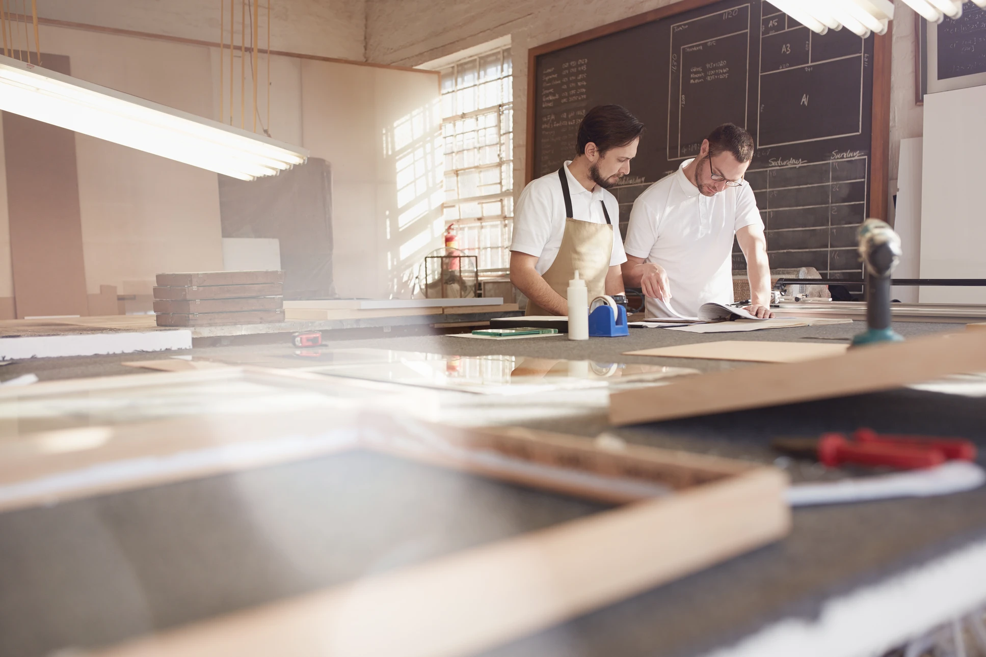 Un homme en chemise verte travaille dans un atelier de menuiserie, parlant au téléphone tout en consultant des documents sur un établi en bois
